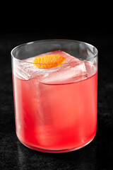 Close-up of a pink cocktail in a clear glass with ice and orange peel garnish on a white...