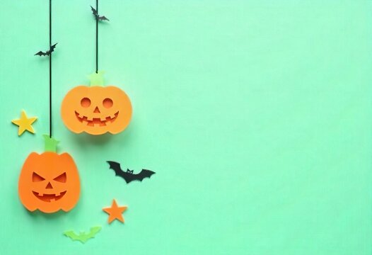 halloween background with space for text. 
Colorful paper cut Halloween decorations hanging from black strings, orange pumpkins with jack-o'-lantern faces, and skulls, against a mint green background