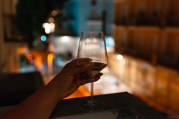 A woman's hand holds a glass of wine on the balcony ledge of a hotel in the summer evening, at night. Vacation time.