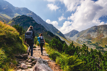 Two women are hiking up a mountain. One of them is wearing a backpack. The other one is carrying a handbag