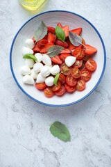 Plate of caprese salad with strawberry, cherry tomatoes, mozzarella cheese and fresh basil, vertical shot on a white stone background, above view