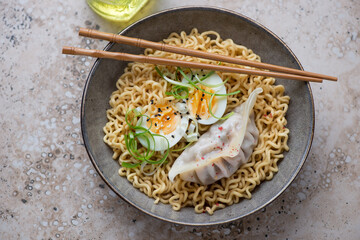Plate of ramen noodles with boiled egg and steamed dumpling on a beige granite background, horizontal shot, elevated view