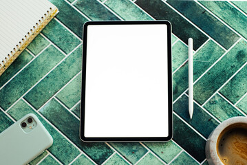 Top view of a digital creative workspace featuring a blank screen tablet, stylus, notebook, smartphone, and a cup of coffee on a stylish green tile surface. Ideal for concepts like remote work