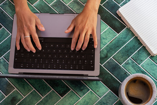 Top view of hands typing on a computer keyboard placed on a green tile surface, with accessories like a notebook, tablet, stylus, smartphone, and a cup of coffee. Represents productivity.