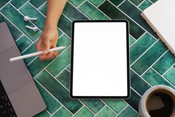 Top view of a woman’s hand holding a stylus above a digital tablet with a blank screen, surrounded by a notebook, coffee, and phone on green tiles. Perfect for creative work, digital drawing