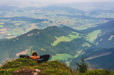 A man is laying on a hillside, looking out over a green valley. The hill is covered in grass and trees