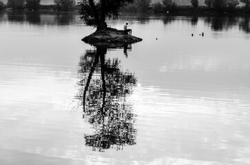 A man sits on a rock in front of a body of water. The water is calm and the reflection of the man and the rock can be seen in the water