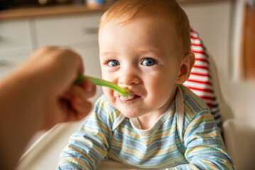little cute boy 8 months old with blond hair and blue eyes eats porridge with a spoon in a high...