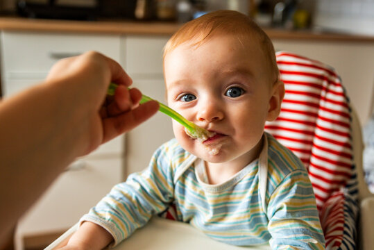 little cute boy 8 months old with blond hair and blue eyes eats porridge with a spoon in a high chair