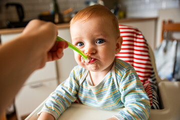 little cute boy 8 months old with blond hair and blue eyes eats porridge with a spoon in a high...