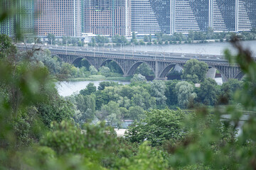 A river bridge spans the water, lined with trees and plants, while contemporary buildings rise in...