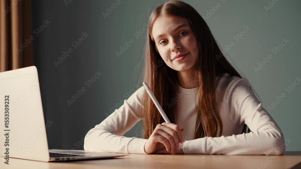 Poster Student girl participating in online class, sitting at desk with laptop, holding pen and smiling confidently while looking directly at camera during virtual learning session