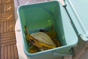 A teal compost bin, open and filled with banana peels and other organic waste, rests outdoors,...