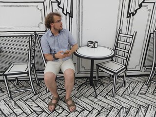 Man is sitting at a table with a cell phone in his hand. There are two chairs around the table...