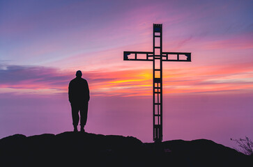 A man stands on a hill with a cross in the background. The sky is purple and the sun is setting