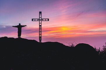 A man is standing on a hill with a cross in the background. The sky is a beautiful mix of pink and purple