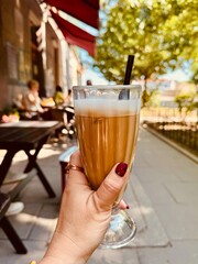 Hand of woman holding a glass of coffee. Latte with milk to drink during break in cafe in centre of town. Close up. Vertical