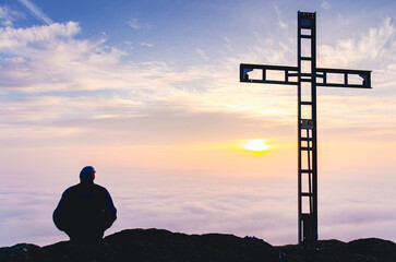 A man is sitting on a hill looking at the sun. The sun is setting and the sky is cloudy