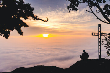 A man sits on a mountain top with a cross in the background. The sky is orange and the clouds are white