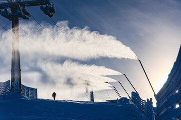 A man is walking on a snowy slope. The snow is falling and the sky is cloudy. There are several ski lifts in the background