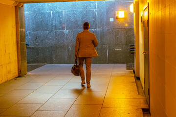 A man walks through an underpass with a brown bag. The hallway is dimly lit. The man is wearing a...