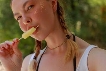 Close-up of a young woman with braided hair and glowing glitter on her cheeks holding and licking a...