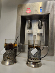 Two cups of tea are sitting on a silver tray next to a water dispenser. The dispenser has a button...