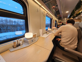 Man is sitting in a train car with a table in front of him. There is a person sitting in the chair...