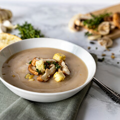 Mushroomsoup with fresh mushrooms, herbs and bread croutons in a white bowl on a marble table with bread in the background