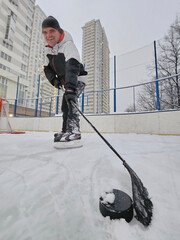 Man wearing a black and white jacket is holding a hockey stick and standing on a frozen lake rink near residential complex