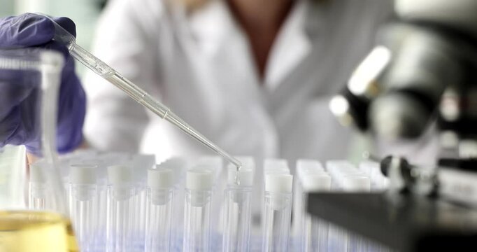 Woman uses pipette to drop liquid reagent into test tubes at experiment in laboratory. Flask and microscope display scientific research and analysis