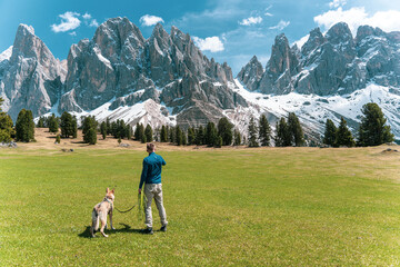 Man with dog standing on green meadow looking at snowy peaks in the Dolomites, Italy.