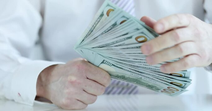 Male office worker in white shirt and tie counts thick stack of hundred dollar bills in office. Financial success and corporate wealth accumulation