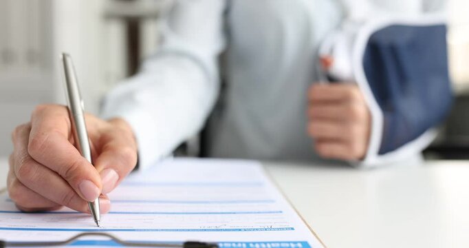 Lady patient with broken arm in sling fills health insurance claim form with silver pen at desk. Concept of healthcare insurance and medical paperwork