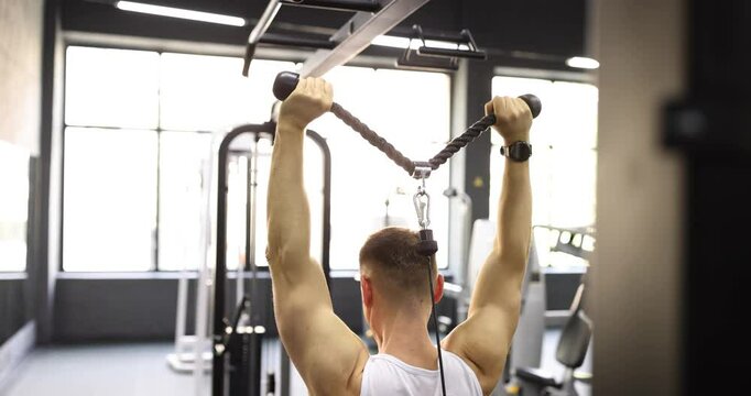 Muscular athlete engages back muscles by pulling down rope attached to weights in modern fitness center. Strength and dedication to physical fitness
