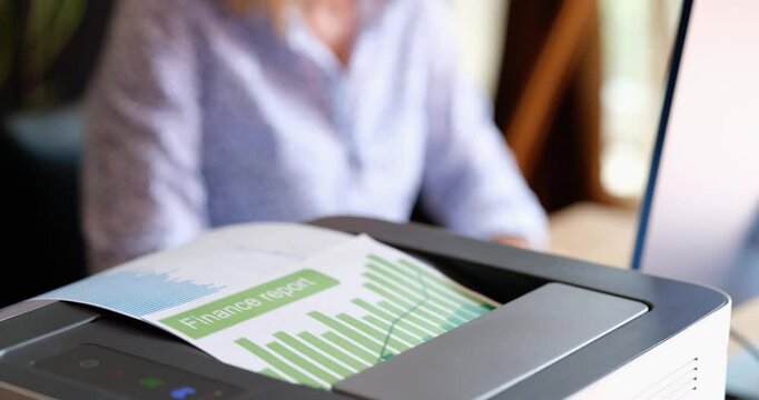 Modern laser printer outputs financial report filled with colorful charts and detailed graphs. Office worker sits at desk and types on keyboard