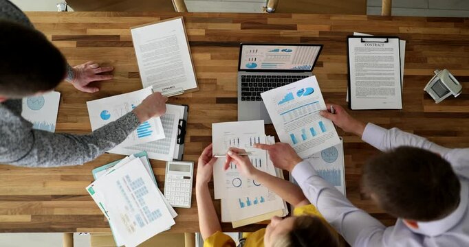 Professional team analyses financial report comparing graphs and data on laptop and papers. Colleagues collaborate at project meeting in modern office