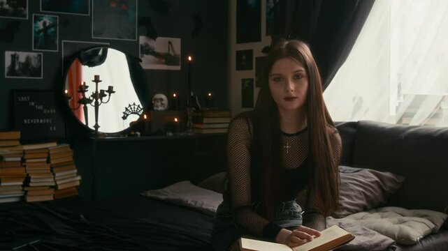 Portrait shot of dark-haired teenage girl in black outfit sits somberly on bed and reading horror book in gothic-themed bedroom