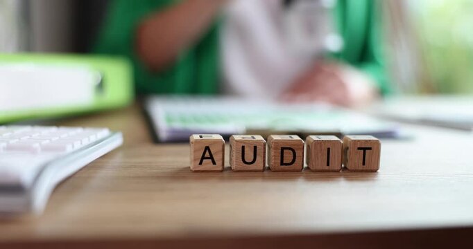 Wooden cubes spell out word Audit on office desk. Businessperson reviews document pages emphasizing financial examination and accounting control