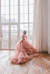 young girl with short black hair in pink princess dress with train is standing from the back and looking cute near huge panoramic white window background at sunny day. fashion concept, free space