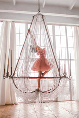 young girl with short black hair in pink princess dress is standing like ballerina on the crystal chandelier near huge white panoramic window background at sunny day. fashion concept, free space