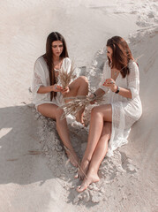 two brunette girls in white boho style dresses sit casual with beige dried flowers in hands and makes a bouquets on the white sand career background. witch cosplay. lifestyle concept, free space