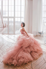 young girl with short black hair in pink princess dress with train is standing from the back and smiling cute near huge panoramic white window background at sunny day. fashion concept, free space