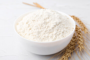 Wheat flour in bowl and spikes on white table, closeup