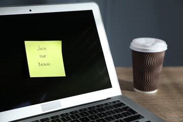Sticky note with words Join Our Team, laptop and takeaway cup on wooden table against grey...