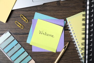 Paper note with word Welcome and stationery on wooden table, flat lay