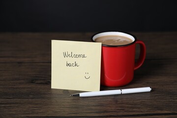 Paper note with phrase Welcome Back, pen and cup of coffee on wooden table