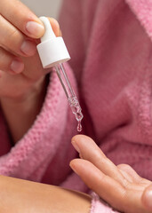 Young woman in pink bathrobe dropping essential oil on a finger and holding a pipette closeup