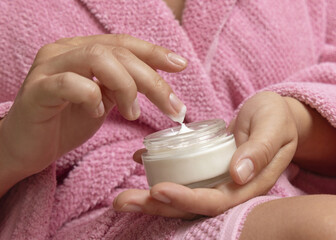 Young woman in pink bathrobe touching cream with finger and holding a cosmetic jar closeup