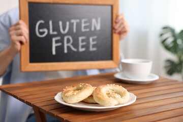 Man holding small chalkboard with text Gluten Free at wooden table indoors, focus on pretzels
